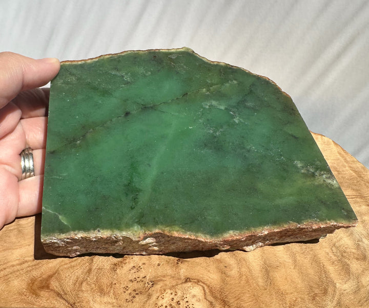 A raw green stone slab with natural marbling and rough edges, held by a hand over a wooden surface against a light gray fabric background.