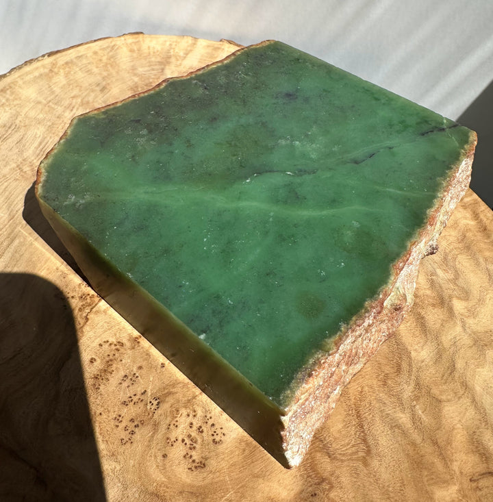 Green jade slab with rough natural edges, resting on a cut wood slice in bright natural light.