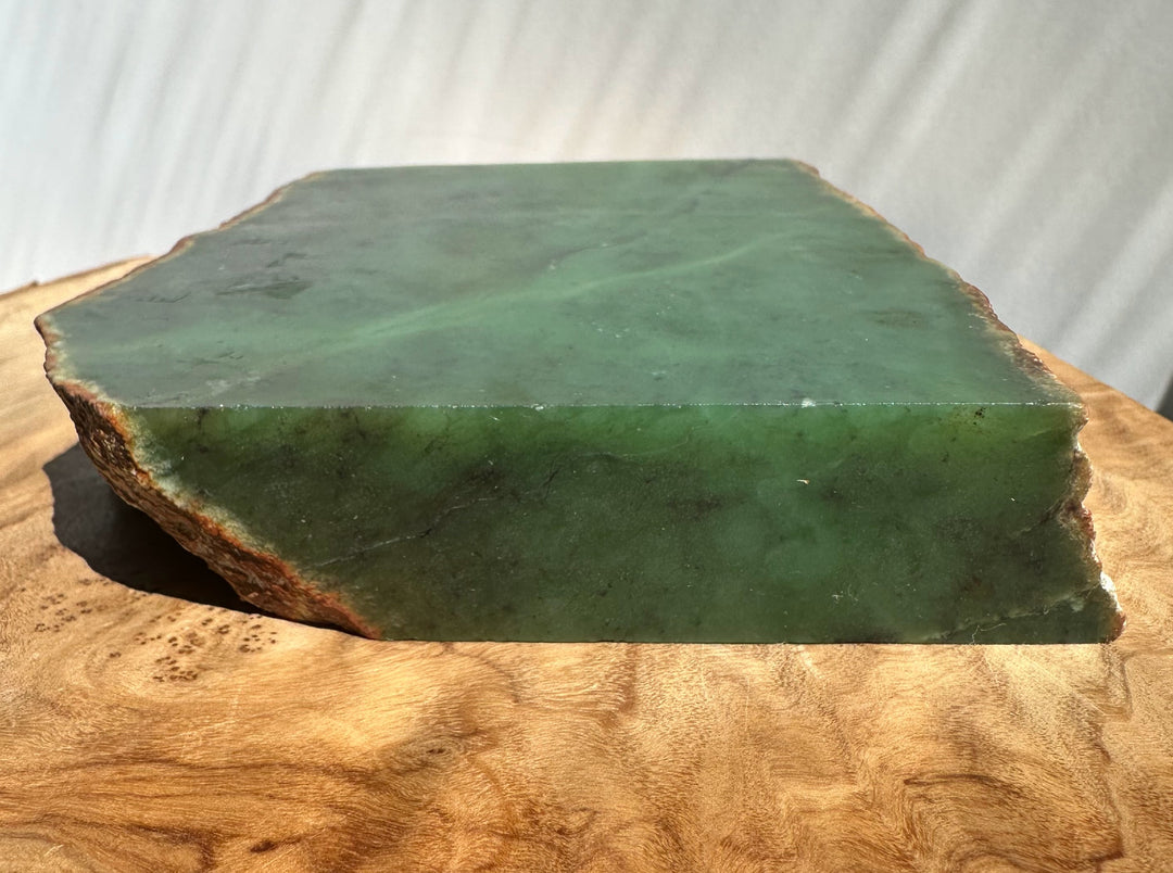 Green jade slab with rough natural edges, resting on a curved wooden surface against a light-draped fabric background.