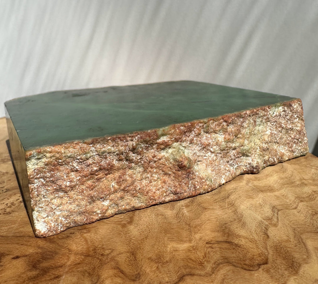 Green jade slab or decorative block with a rough, textured brown and beige natural edge, displayed on a curved wooden surface against a light gray draped backdrop.