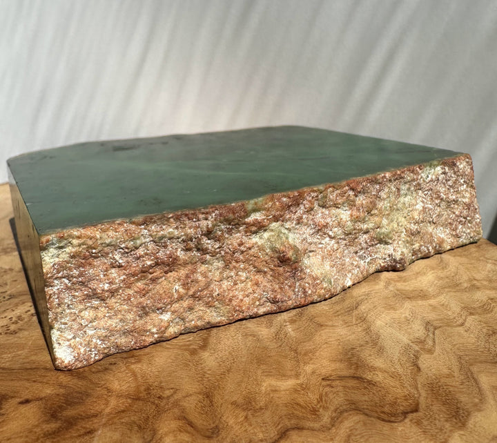 Green jade slab or decorative block with a rough, textured brown and beige natural edge, displayed on a curved wooden surface against a light gray draped backdrop.