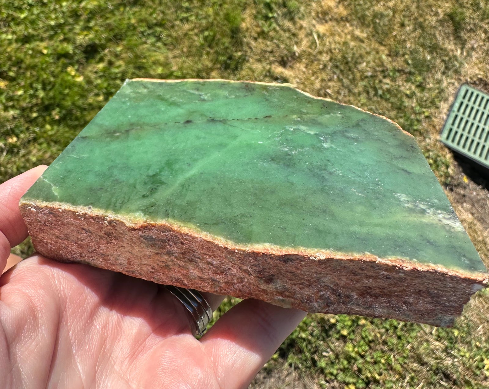A hand holding a raw green jade slab with a rough, brown-textured edge, photographed outdoors on grass.