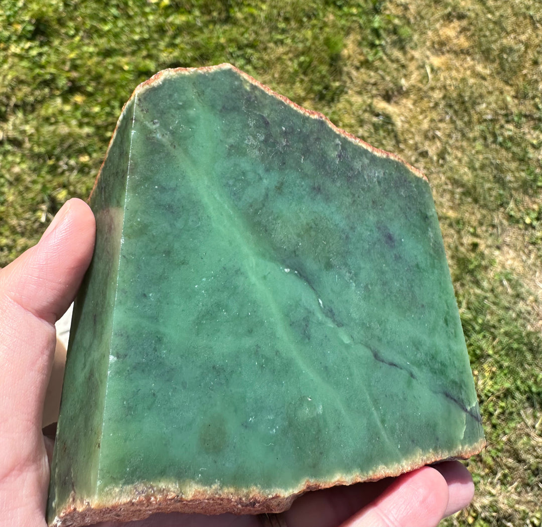 A hand holding a large, rough green jade slab with natural marbling and brownish edges, set against a blurry background of grass.