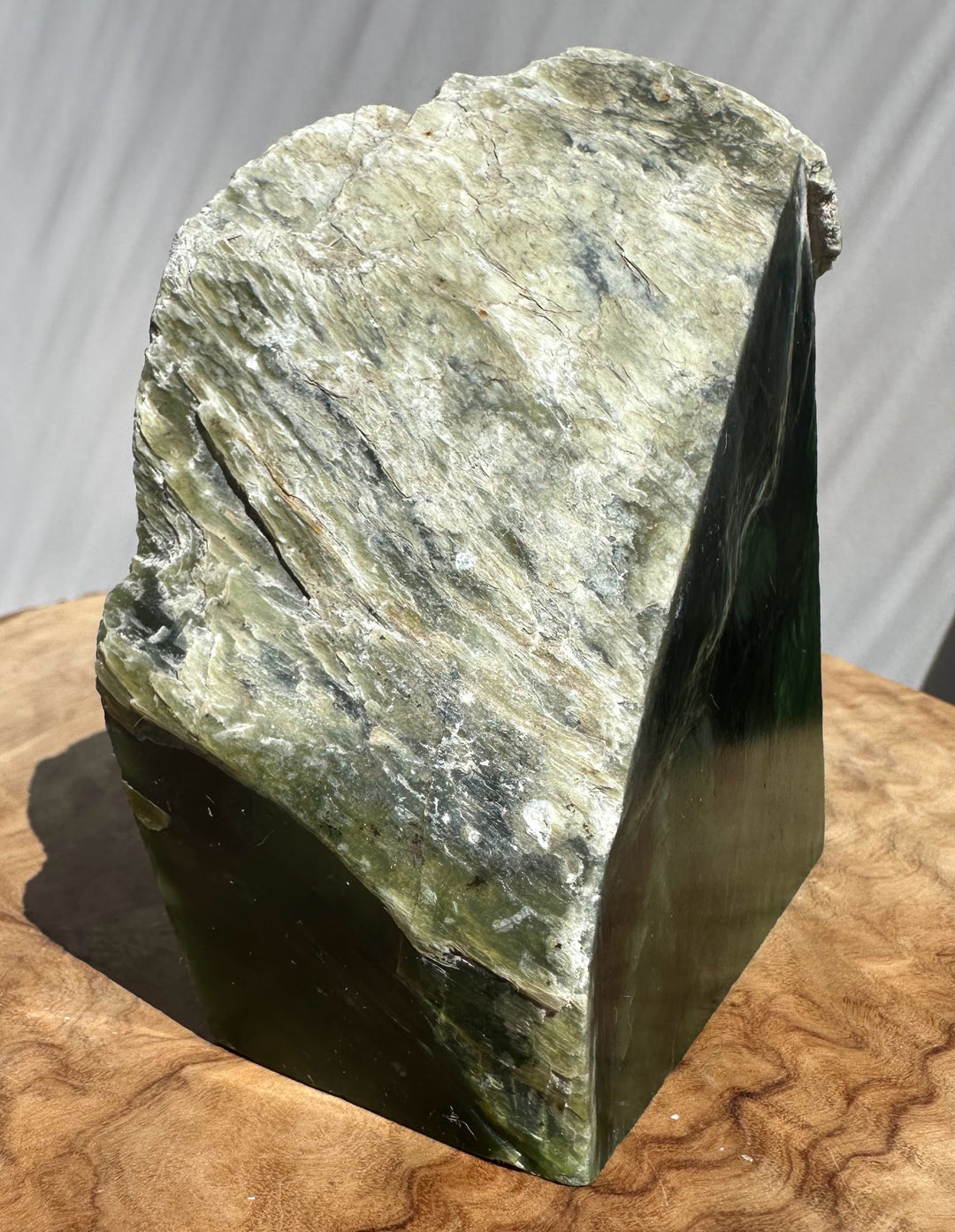 Natural jade stone sculpture with a wavy, layered top section in olive green, gray, and cream, resting on a curved wooden surface against a light gray draped backdrop.