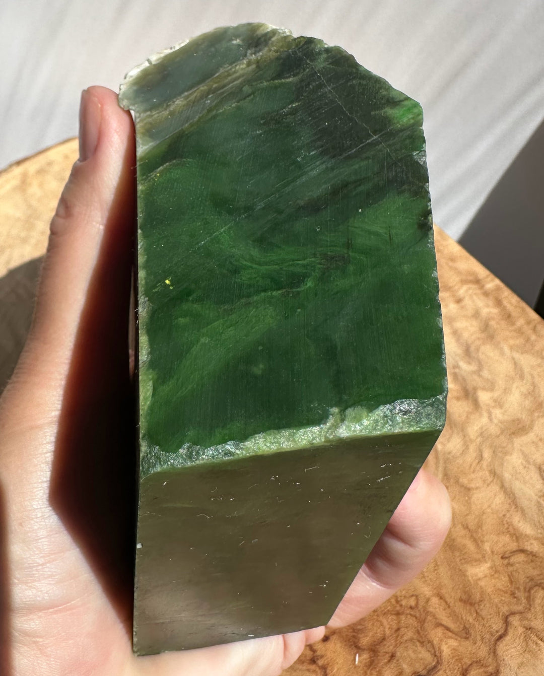 A hand holding a green jade rectangular block with a natural rough edge, photographed against a wooden surface and light background.