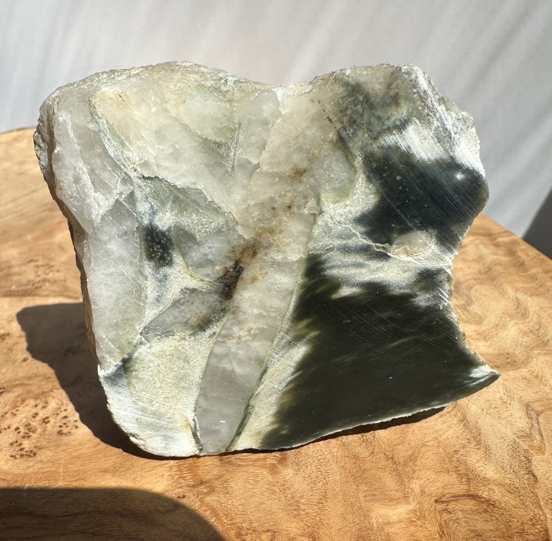 A raw stone or mineral slab with natural fissures, shown standing upright on a wooden surface. The stone has a mix of light gray, off-white, and dark charcoal/black areas, with a glossy finish.