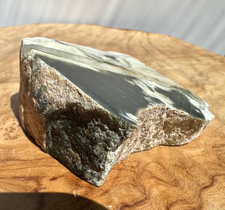 A raw jade with a smooth dark gray to black top surface and a rough brown textured side, displayed on a wooden surface.