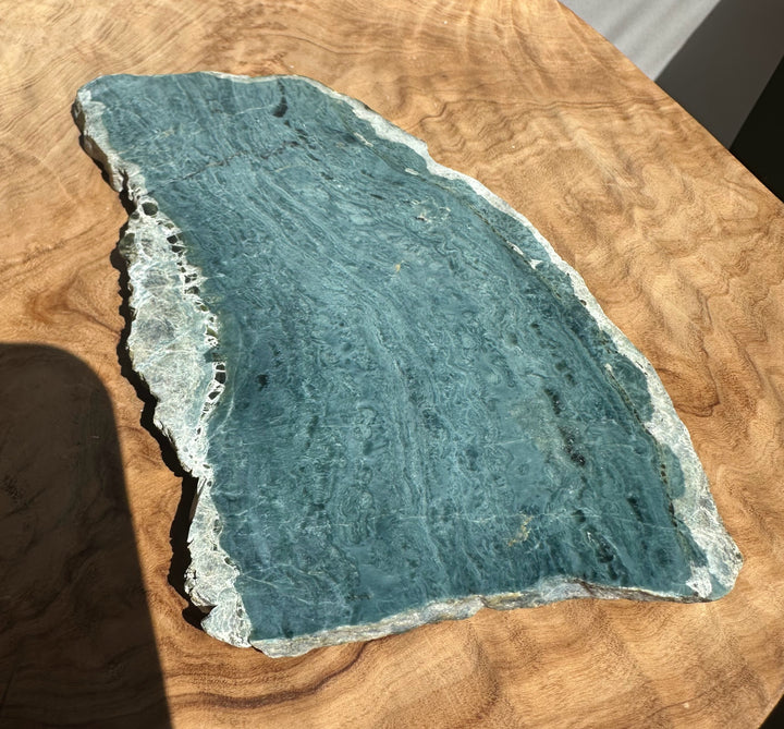 A raw, blue-green stone slab with natural marbling and a rough, uneven edge, displayed on a warm-toned wooden surface.