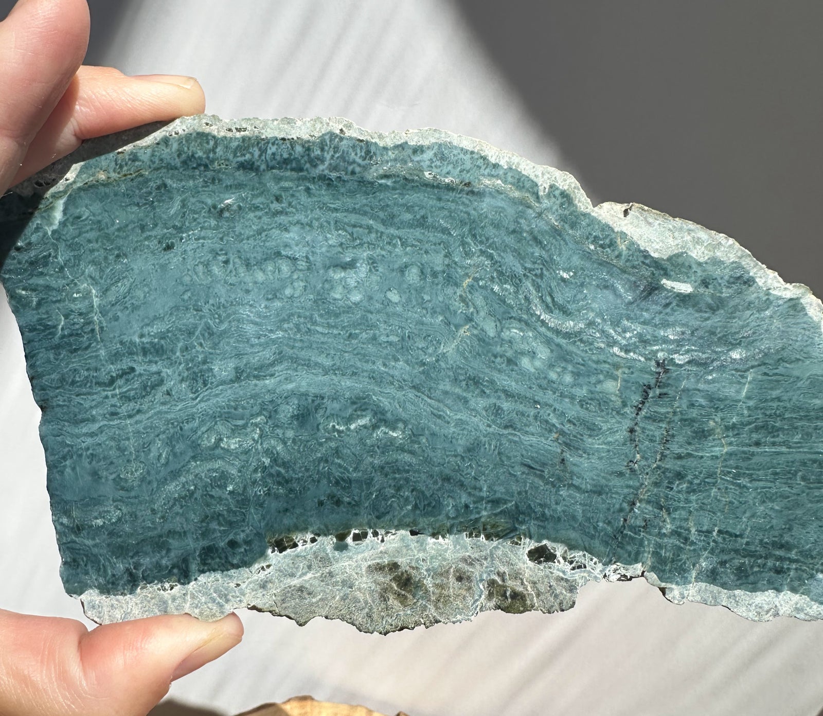 A hand holding a raw, irregularly shaped slab of blue-green stone or mineral with visible natural texture and layered patterns.