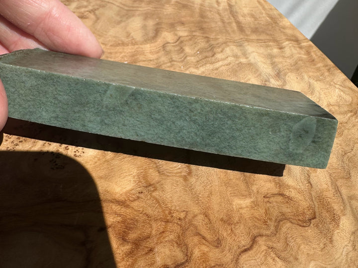 A rectangular green jade-like block held by a hand over a highly polished burl wood surface.