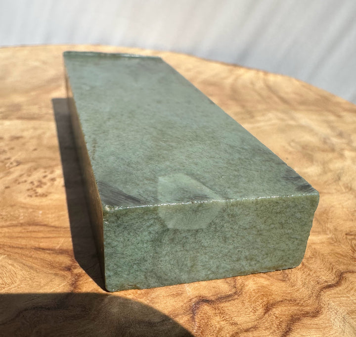 A rectangular green stone, jade, resting on a round wooden surface with a white draped background.