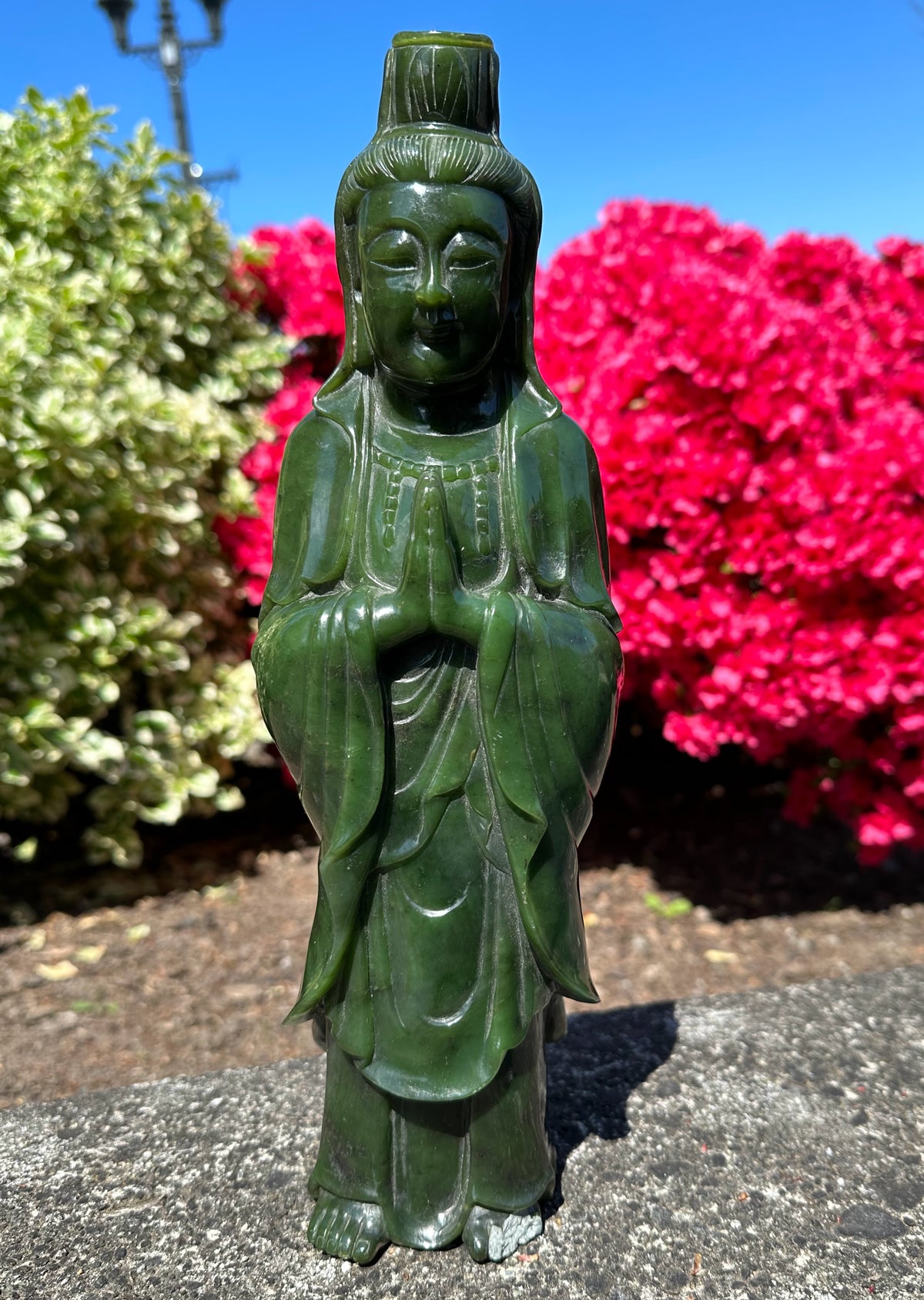 12.5 inch dark green Canadian jade Guanyin statue in a prayer pose outdoors with a blue sky and red flowers behind