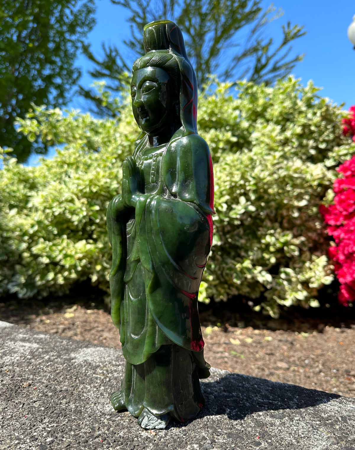 Side view of a dark green Canadian jade Guanyin statue in a praying pose outdoors among greenery and red blossoms
