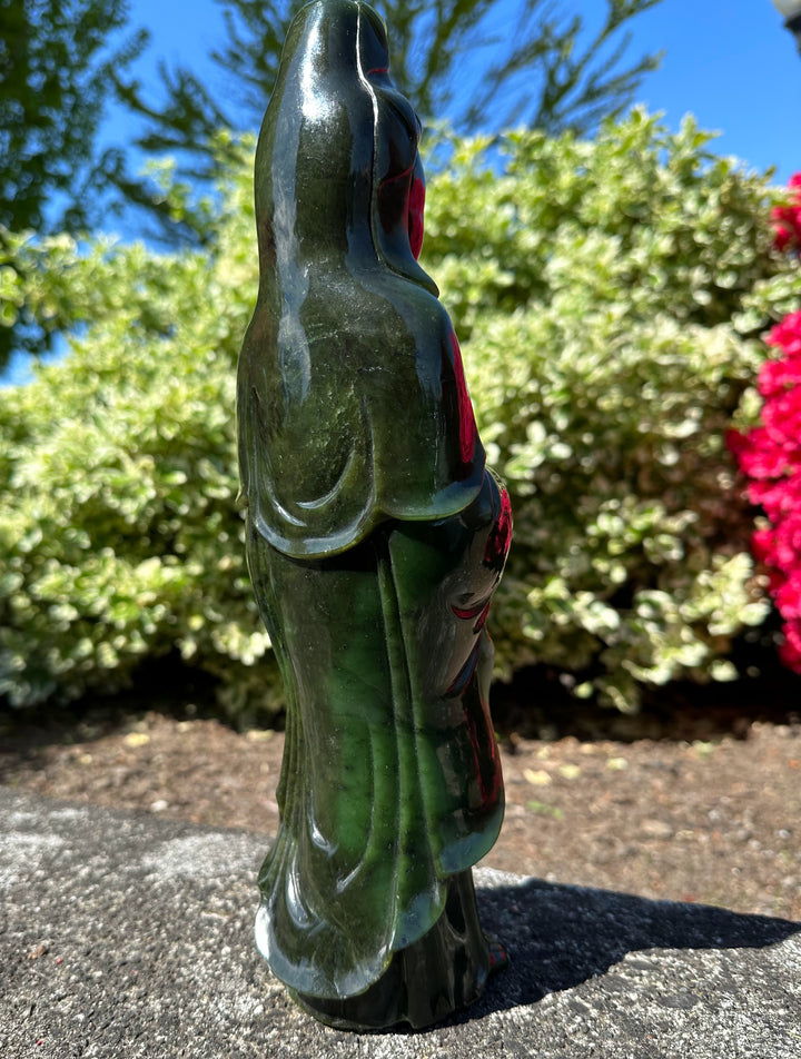 Tall dark green Jade Guanyin statue outdoors on a stone surface with greenery in the background