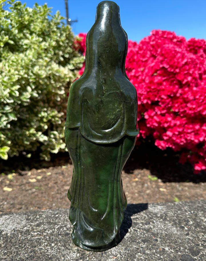 Green jade Guanyin statue standing outdoors with pink flowering bushes in the background.