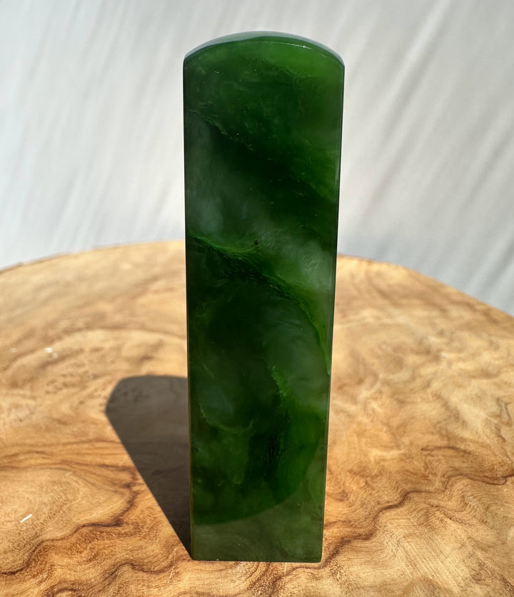 A polished green jade rectangular block with rounded top corners, standing upright on a rustic wood slice surface.