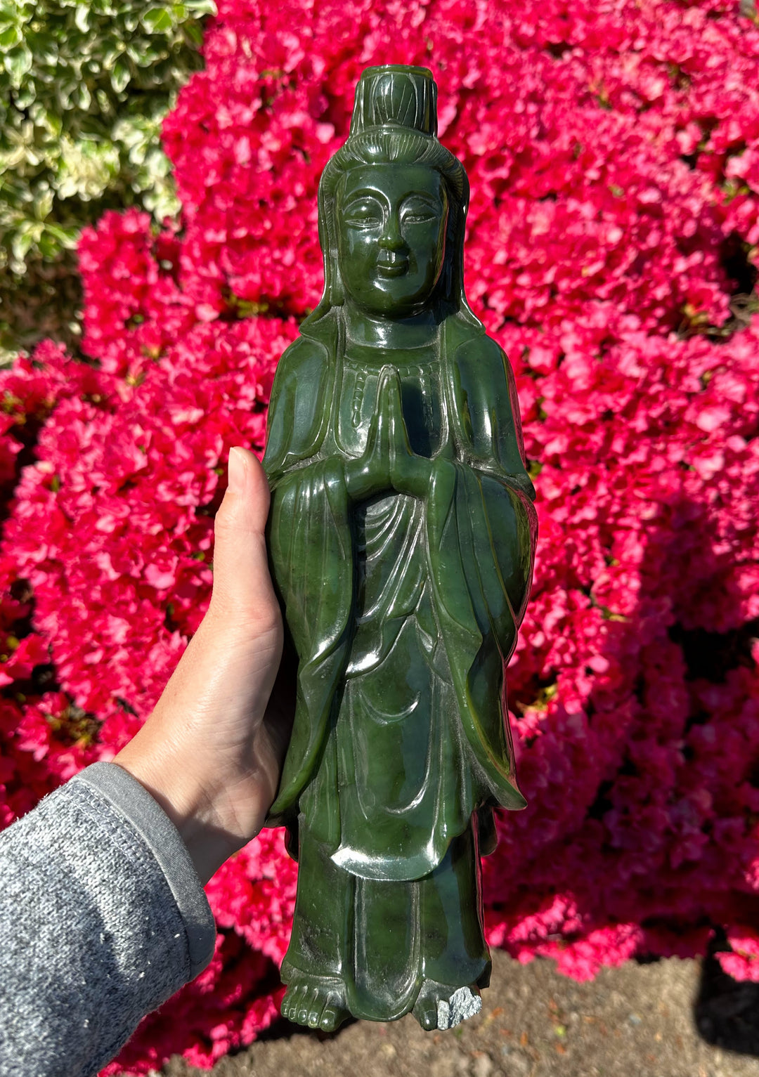 Green Canadian jade Guanyin statue held in a hand with pink blossoms behind.