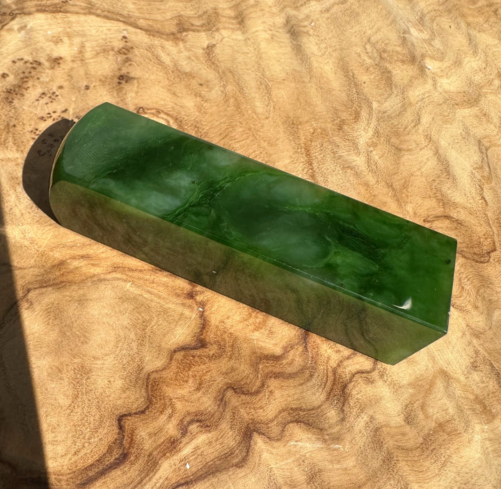 A polished rectangular green jade rounded ends, resting diagonally on a textured wavy wood surface.