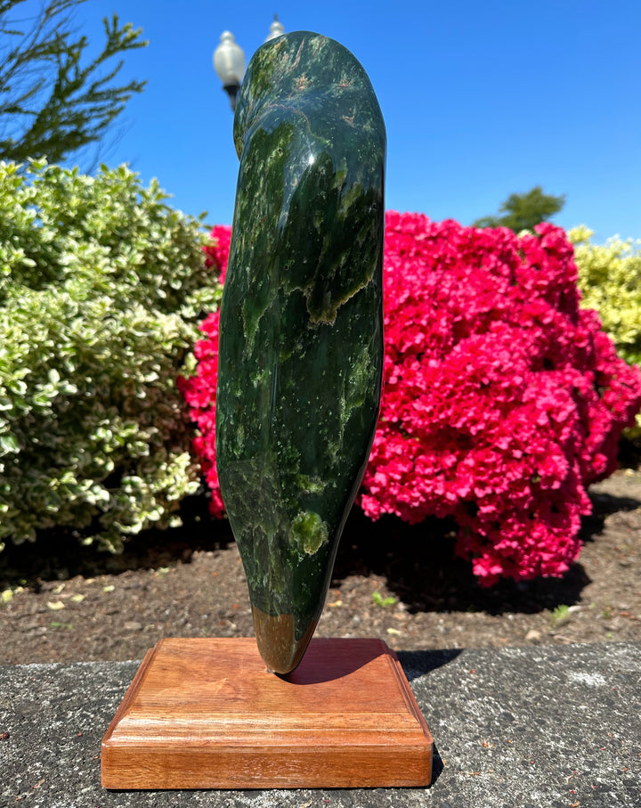 Tall polished Canadian jade obelisk on a wooden base outdoors with blue sky and bright pink azaleas.