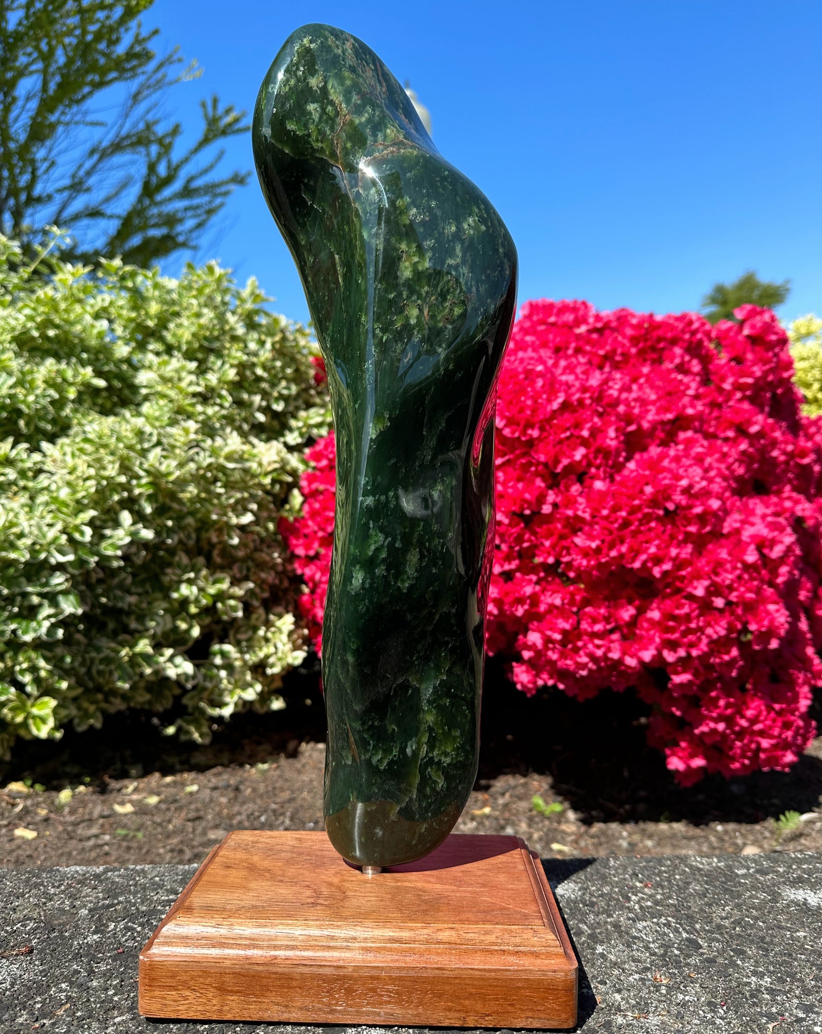 Polished Canadian jade sculpture with a curvy abstract form on a wooden base outdoors beside pink flowering shrubs.