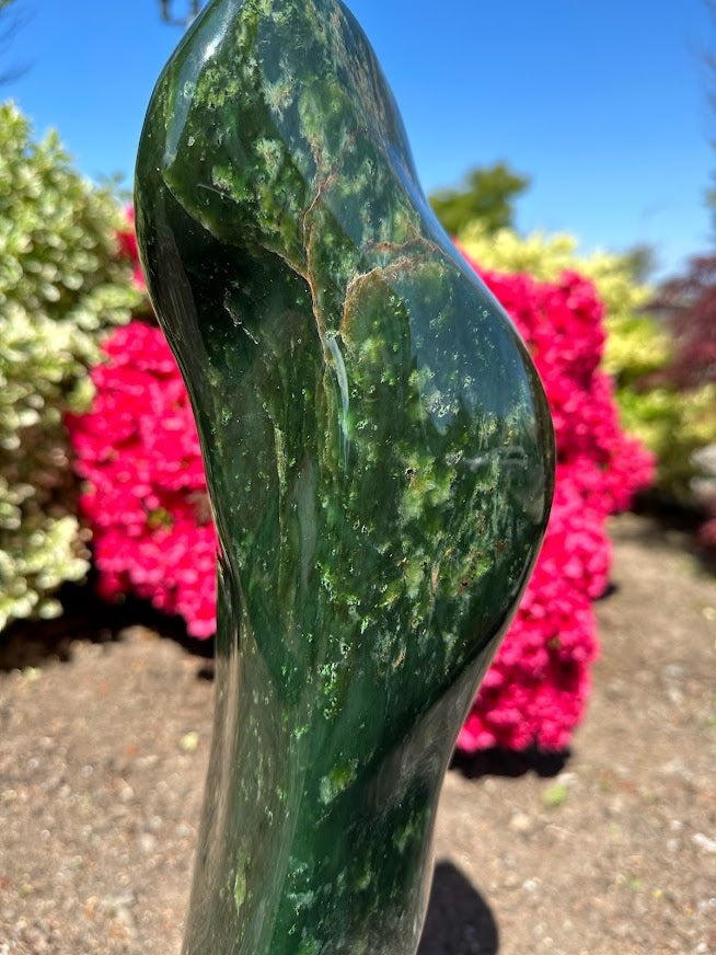 Tall polished Canadian jade sculpture viewed from the side with a small price sticker, mounted on a wooden base outdoors.