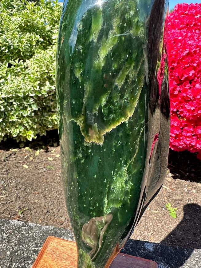 Close up side view of a tall polished Canadian jade specimen on a wooden base outdoors with greenery and pink flowers.