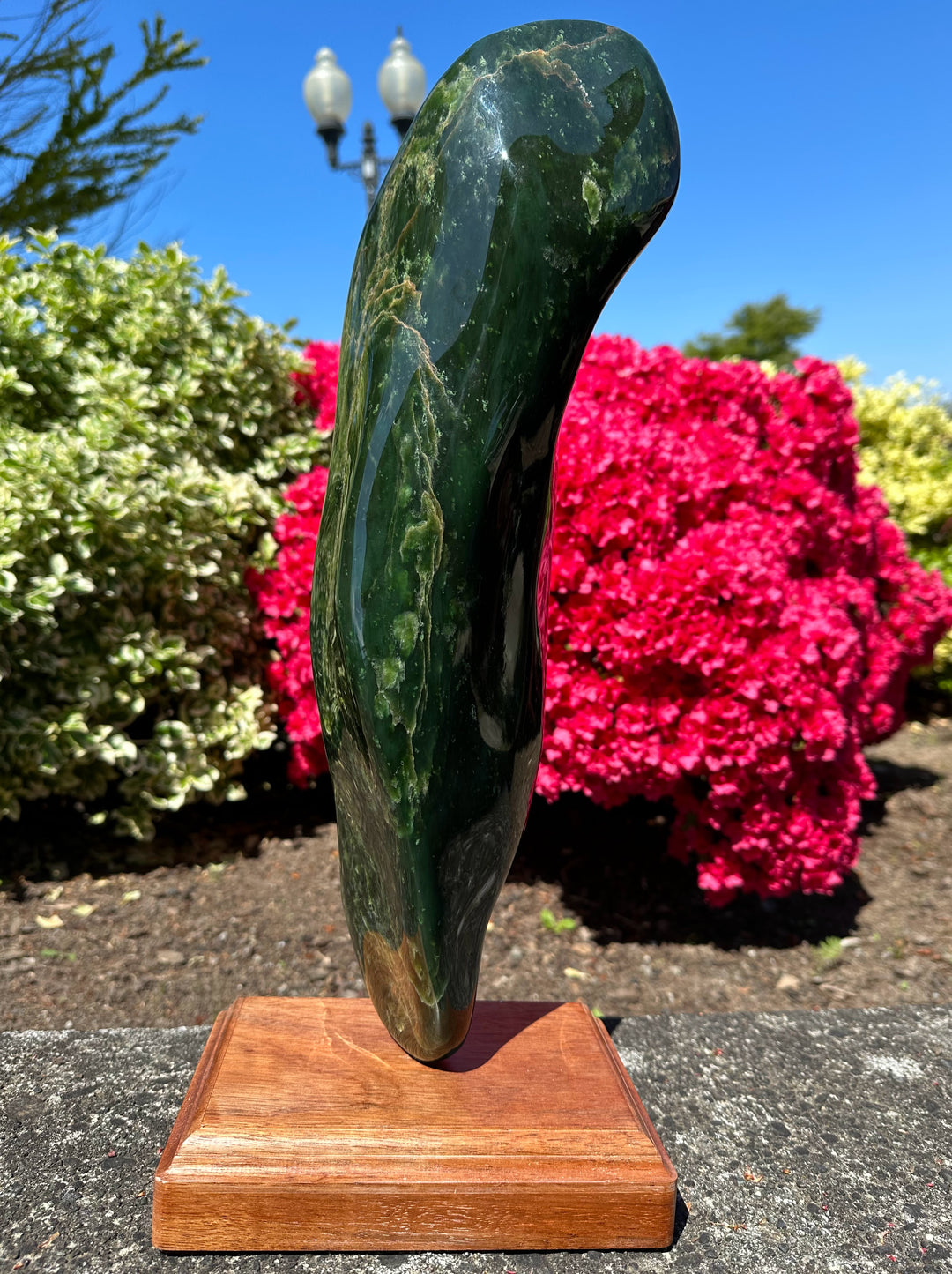 18 lbs 6 oz polished Canadian jade specimen mounted on a wooden pedestal outdoors with blue sky and pink azaleas.