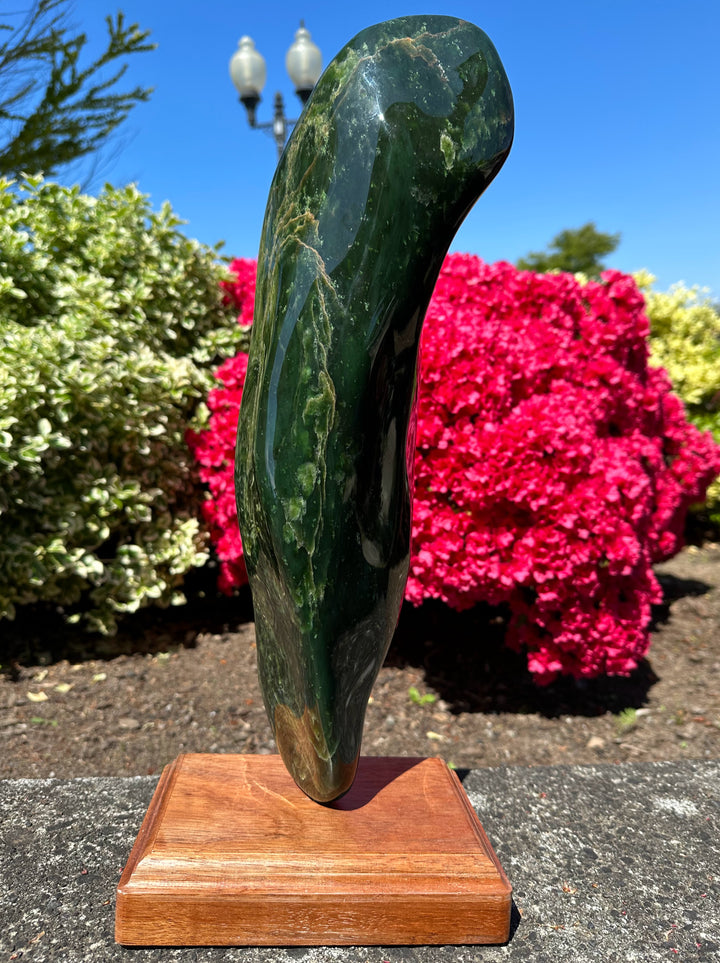 18 lbs 6 oz polished Canadian jade specimen mounted on a wooden pedestal outdoors with blue sky and pink azaleas.