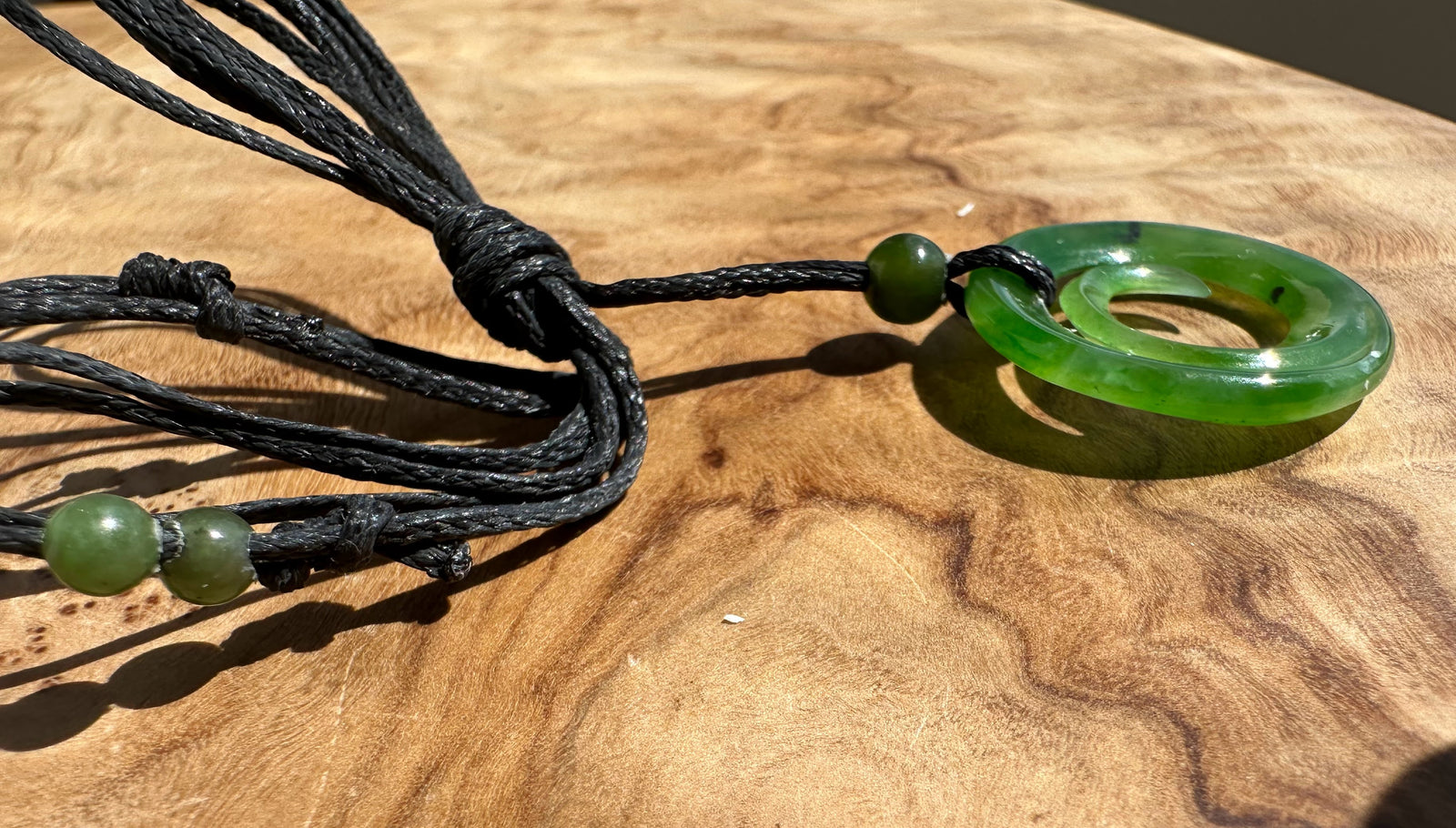 A green jade pendant necklace with a black multi-strand cord and green accent beads, displayed on a wooden surface.
