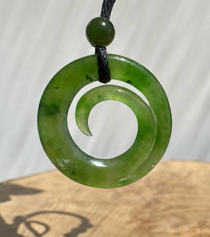 A green polished stone spiral pendant necklace with a matching round bead on a black cord, shown against a light neutral background with a wooden surface below.
