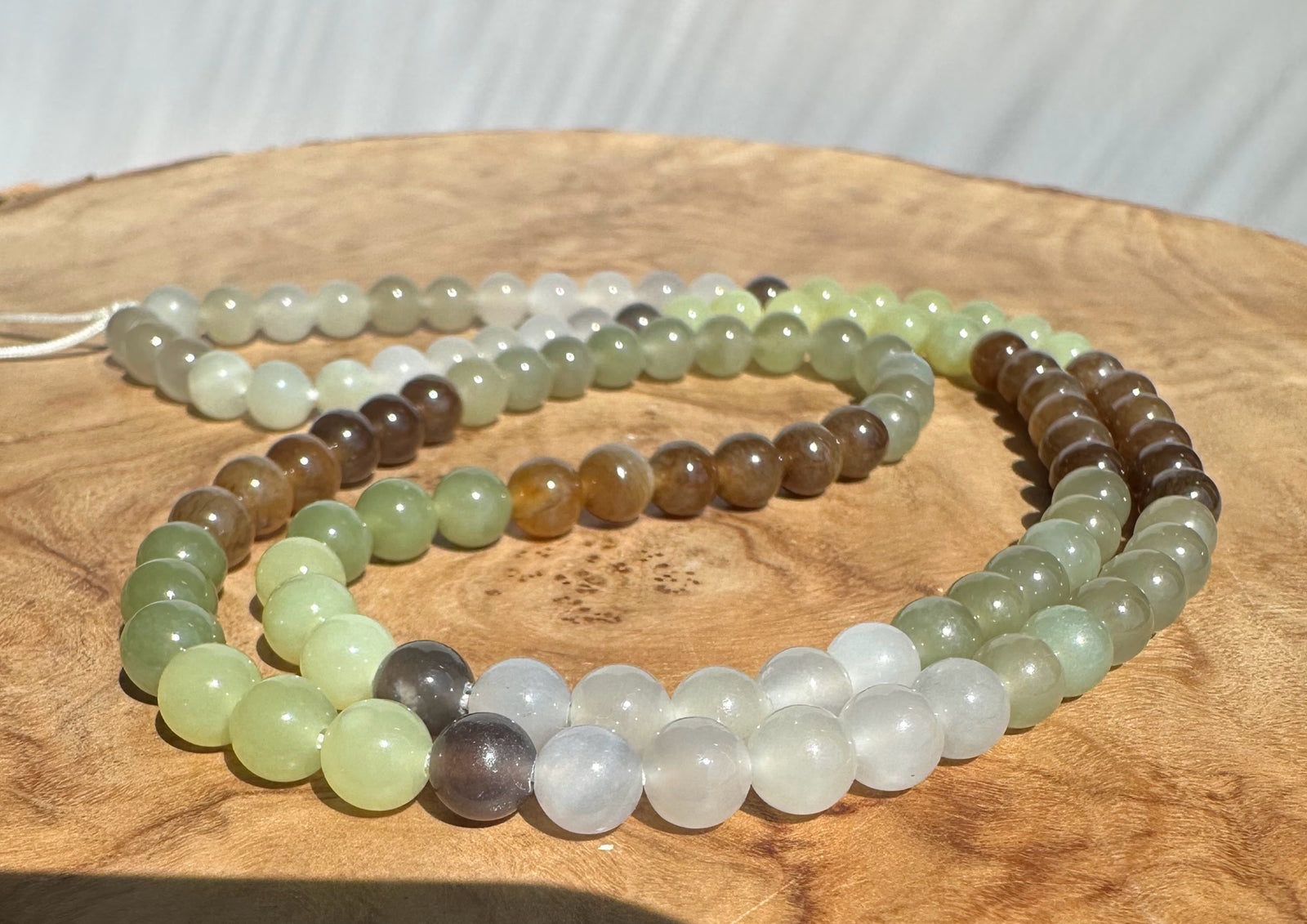 A strand of polished round gemstone beads in soft green, white, brown, and gray tones, arranged in loops on a round wooden surface.