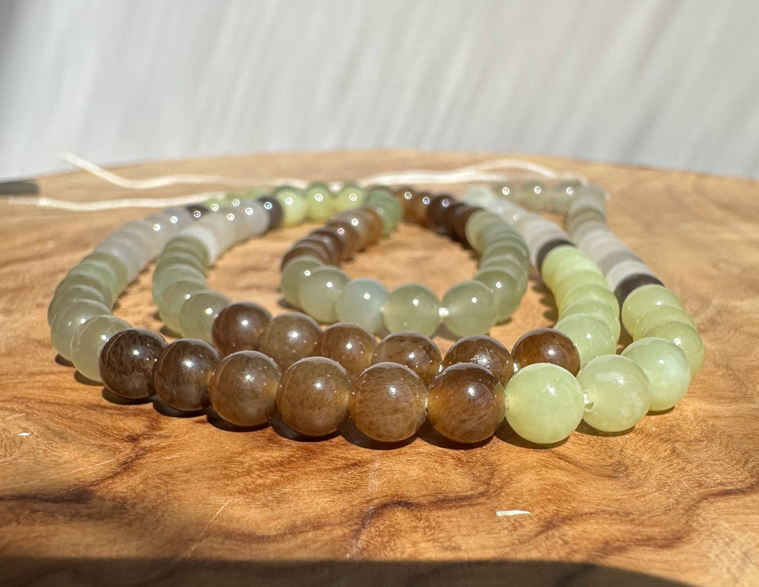 Unstrung round jade beads in earthy tones, arranged on a wooden surface. The necklaces include shades of brown, olive green, pale yellow-green, and gray.