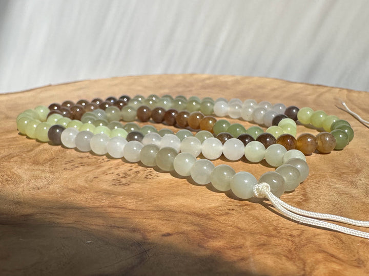 A set of multi-strand beaded prayer beads or mala necklaces made from polished natural stones in various earthy shades of green, brown, gray, and white, resting on a rustic wood slice with a white cord threaded through.