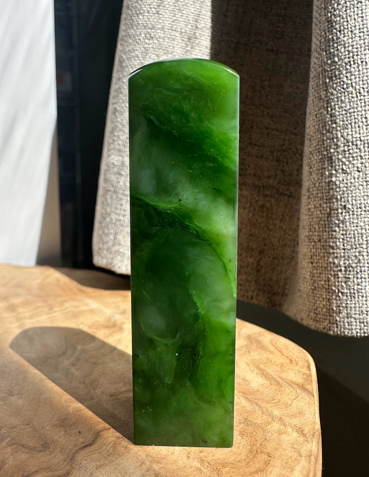A polished green jade rectangular block with rounded top corners, standing upright on a wooden surface with draped fabric in the background.