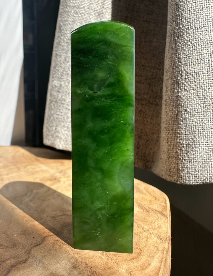 A polished rectangular green jade stone block with rounded top corners, standing upright on a wooden surface with a textured neutral fabric backdrop.