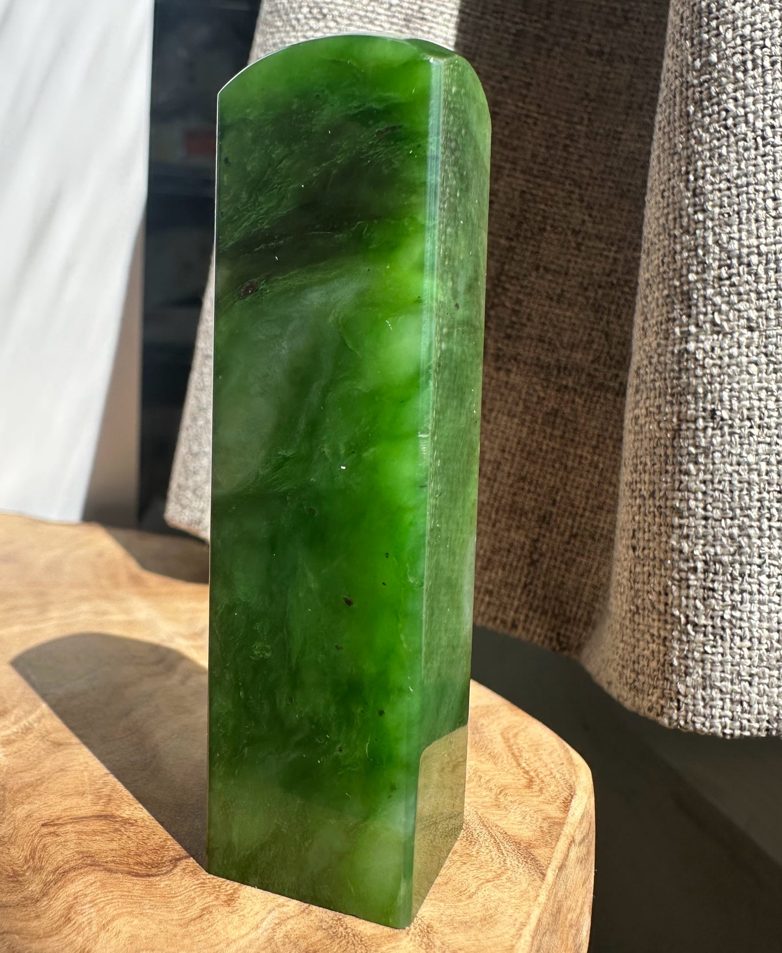 A polished green jade rectangular block with rounded top corners, standing upright on a wooden surface with a textured fabric backdrop.