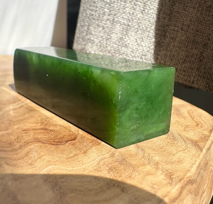 A polished rectangular green jade stone block resting on a wooden surface with a textured fabric background.