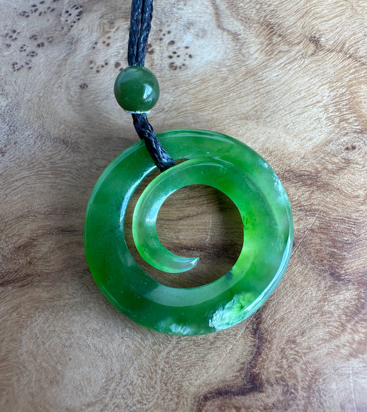 A green stone pendant necklace with a spiral cutout and a round green bead on a black cord, displayed on a textured wooden surface.