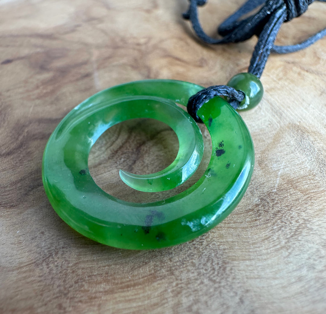 A green polished stone pendant with a spiral cutout, attached to a black cord, resting on a wooden surface.