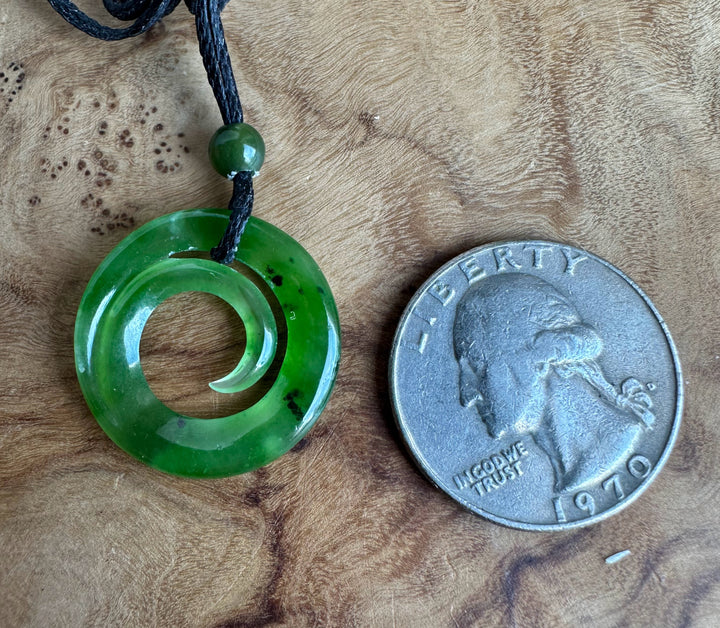 A green carved pendant necklace with a black cord and a green bead, shown next to a 1970 US quarter for scale on a textured wood background.