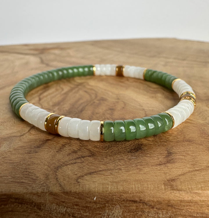 A beaded stretch bracelet featuring alternating green and white cylindrical beads, accented with small gold spacer rings, displayed on a wooden surface.