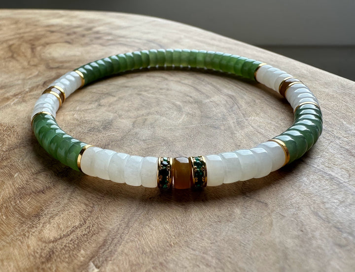 A beaded stretch bracelet with green faceted beads, white opaque beads, and gold-toned spacer rings, displayed on a wooden surface.