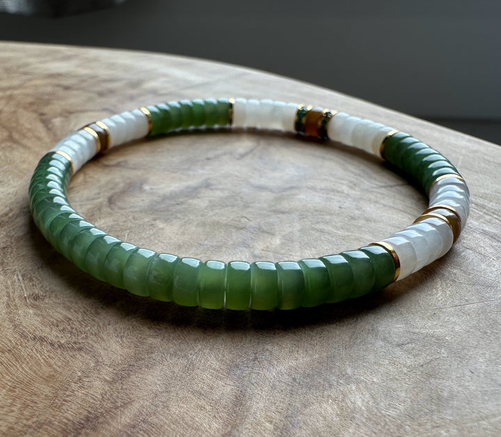 A beaded stretch bracelet featuring faceted green beads and smooth white beads, separated by small gold-toned spacer rings, displayed on a textured wooden surface.