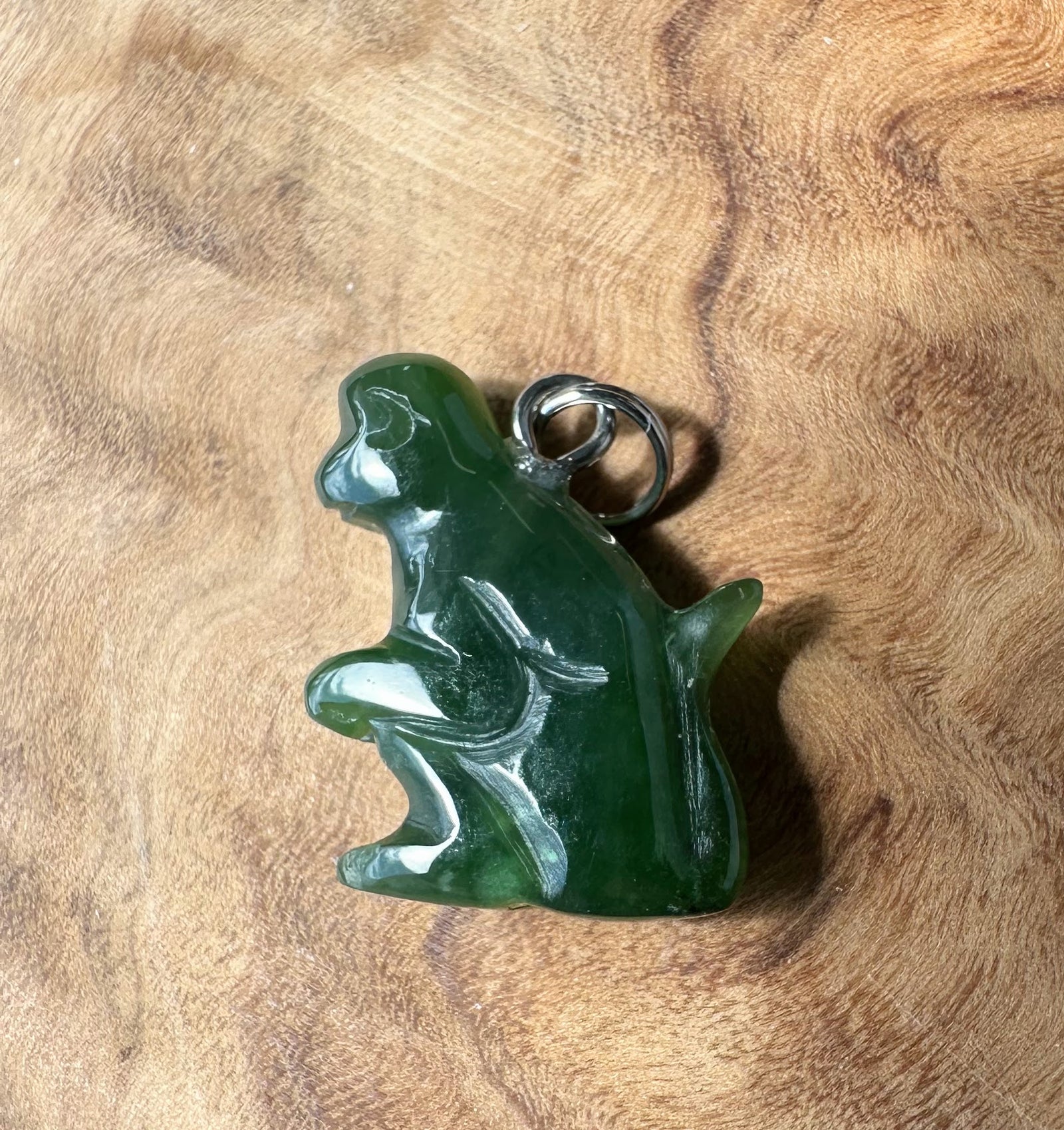 A carved green jade monkey pendant with a silver-colored metal loop at the top, displayed on a textured wooden surface.
