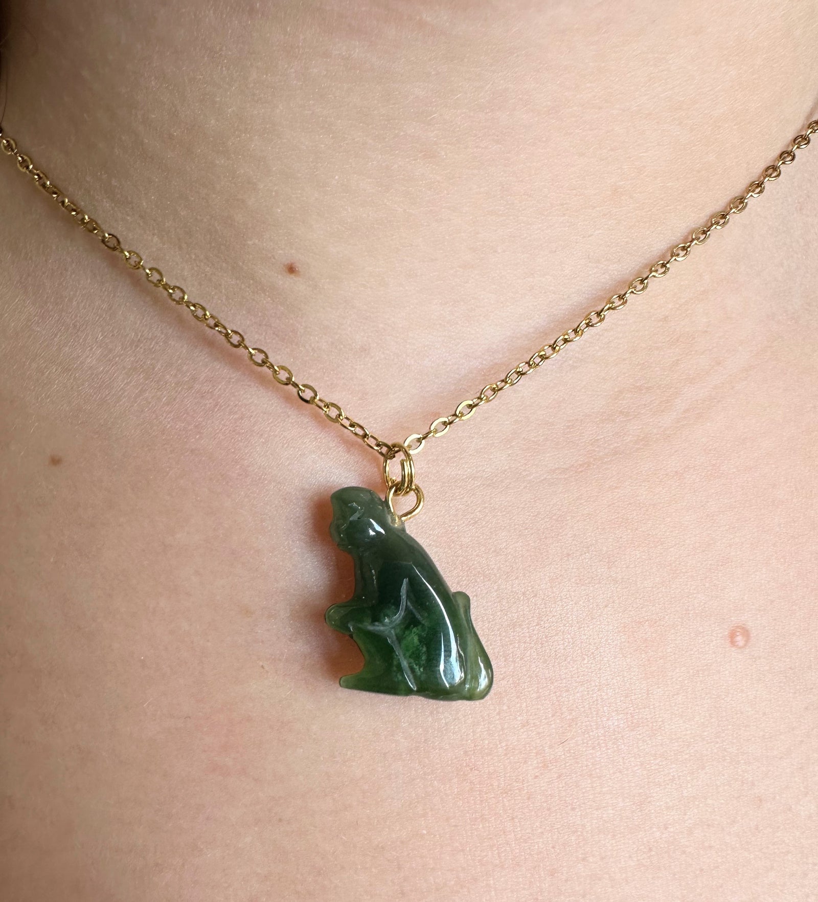 A close-up of a person's neck wearing a delicate gold chain necklace with a carved green jade monkey pendant.