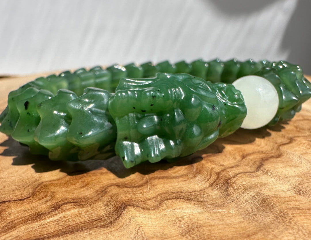 A carved green jade bracelet with a detailed dragon head motif and a smooth white round bead, displayed on a wooden surface.