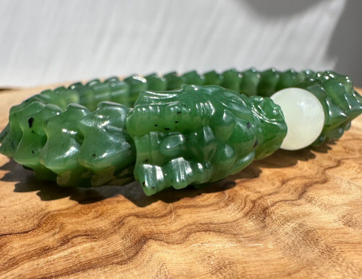 A carved green jade bracelet with a detailed dragon head motif and a smooth white round bead, displayed on a wooden surface.