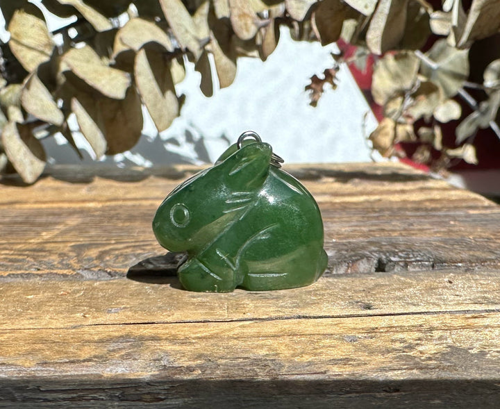 Green bunny shaped nephrite jade pendant with a metal loop bail on a rustic wooden surface.