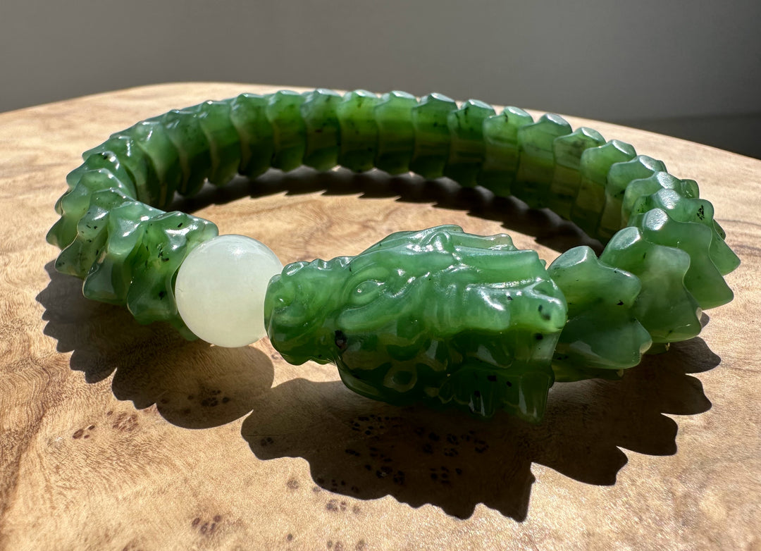 A carved green jade bracelet with a detailed dragon head centerpiece and a smooth white bead, displayed on a wooden surface.