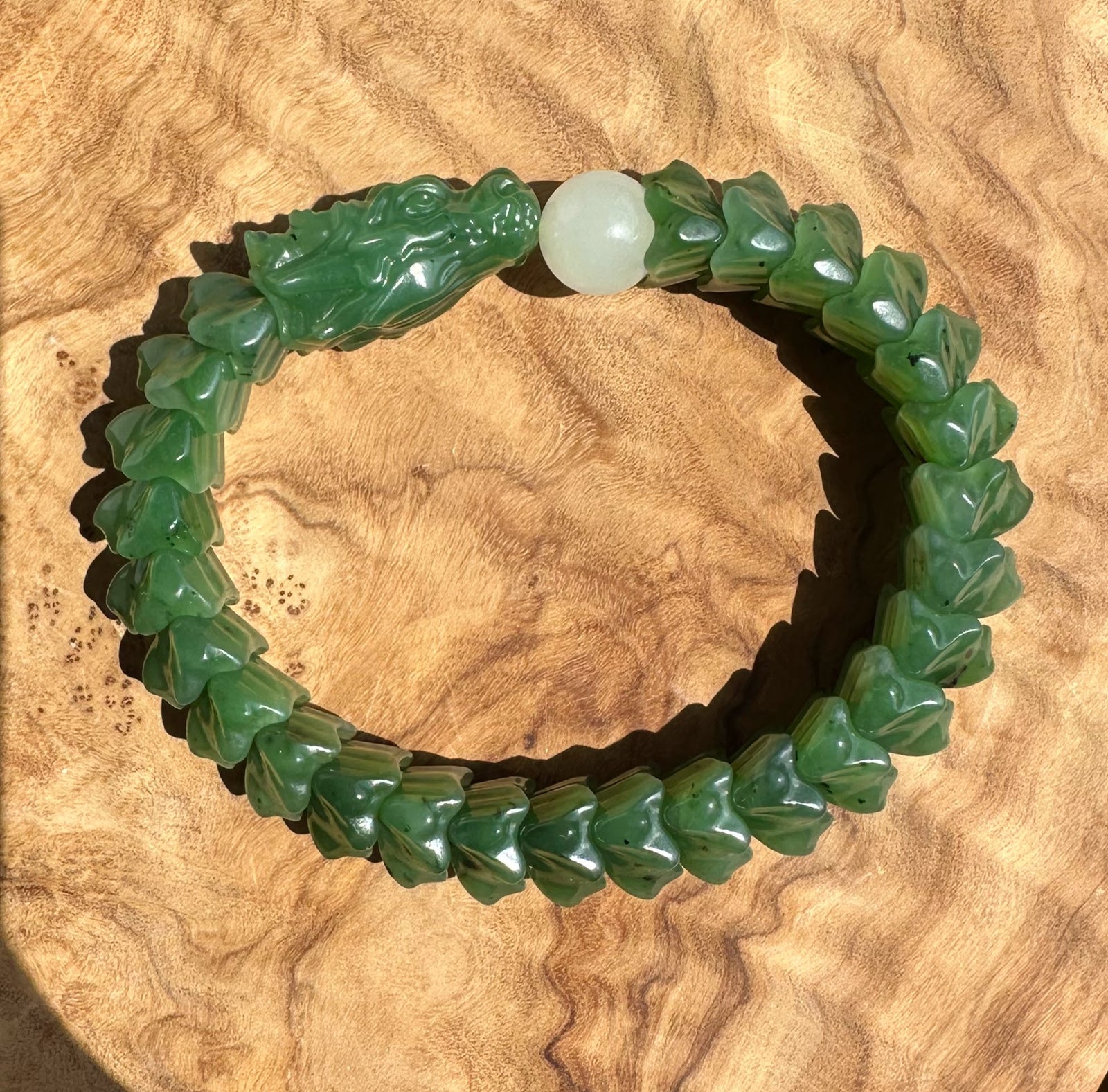 A green jade-like beaded bracelet with a carved green dragon head charm and a round white bead, displayed on a textured wooden surface.
